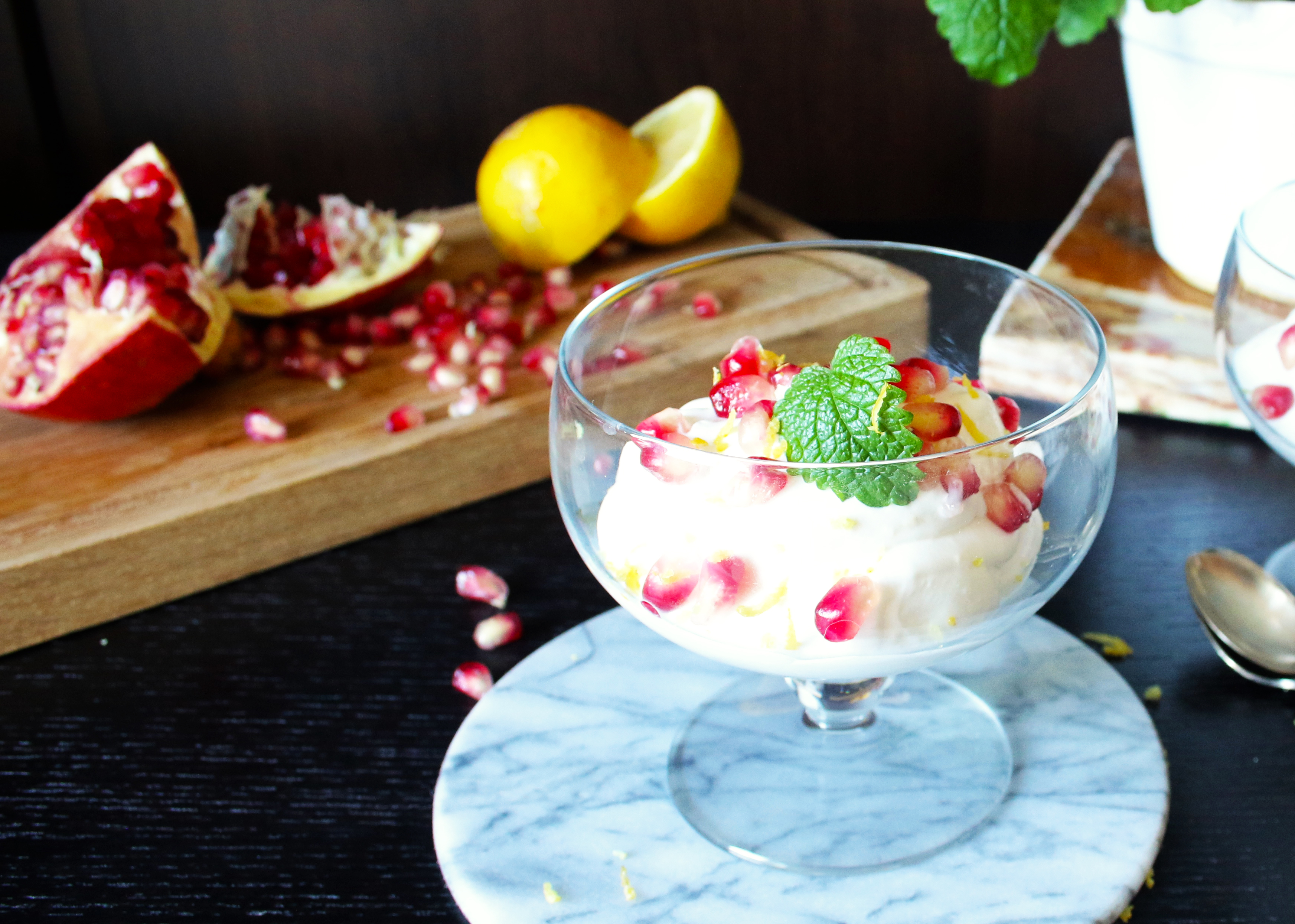 Glasskål med citronmousse och granatäppelkärnor framför en träskiva med granatäpplen och citroner.