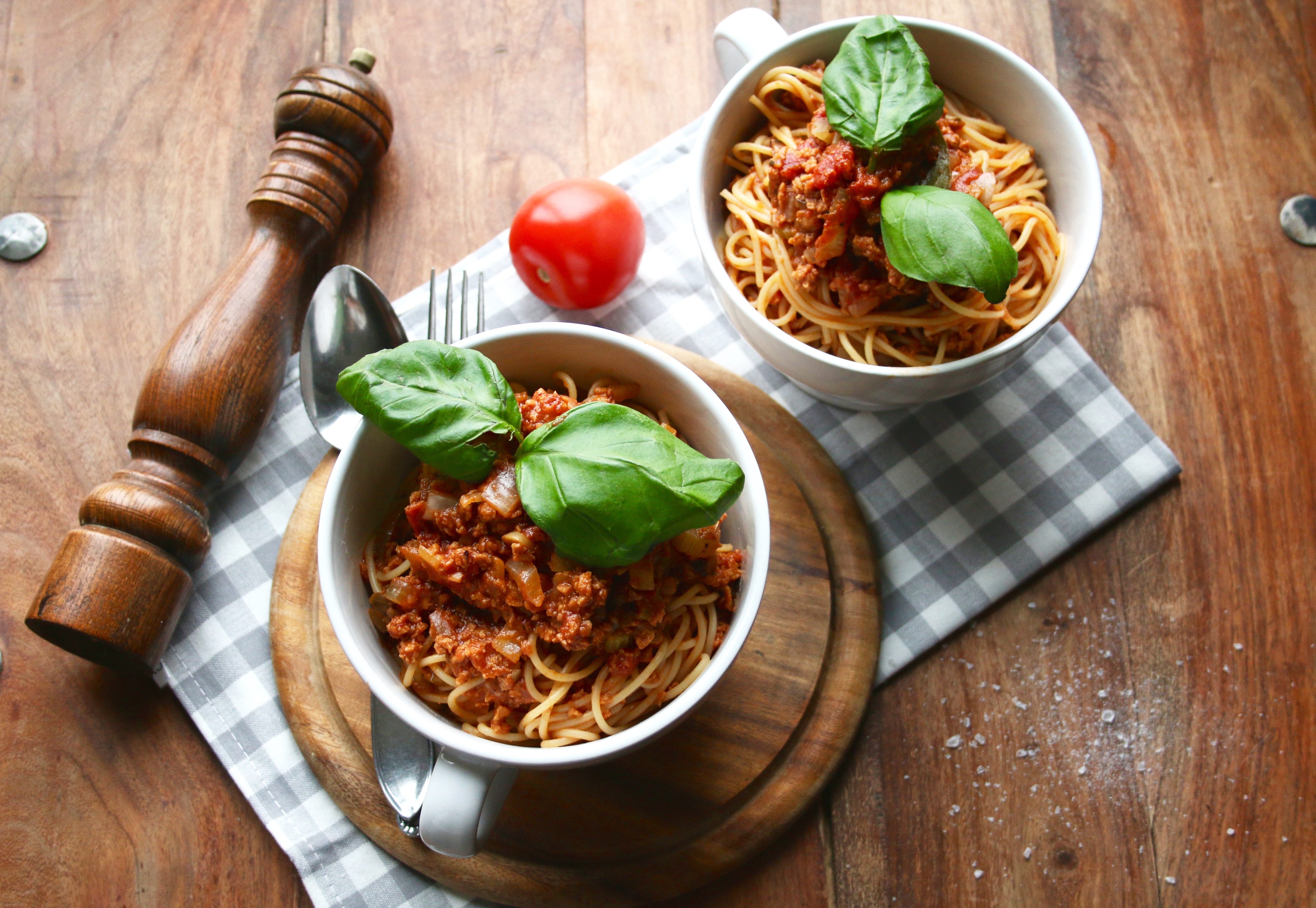 Två skålar med pasta bolognese på ett träbord. Det ligger en pepparkvarn och en tomat brevid skålarna.