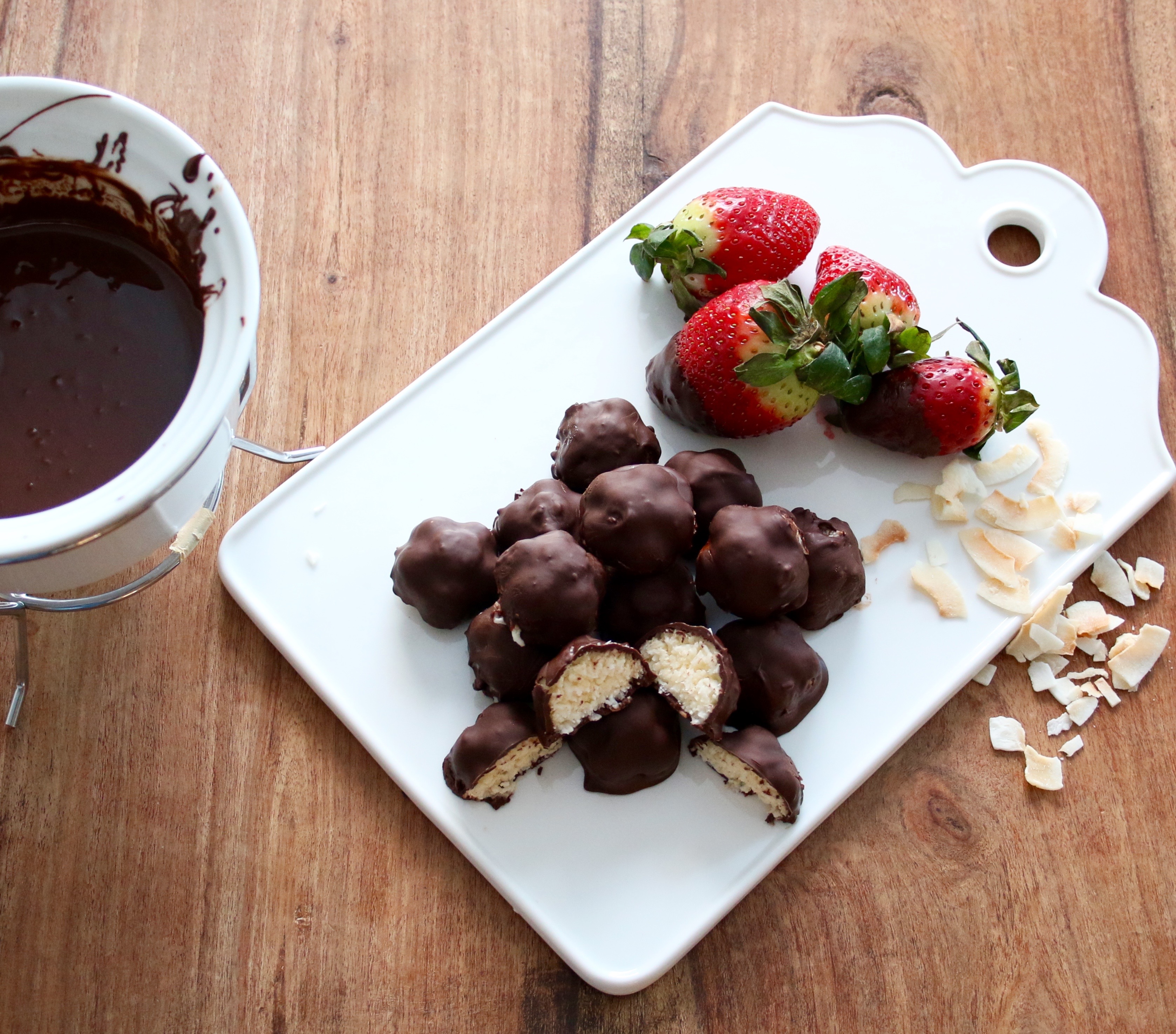 Chokladbitar med kokosfyllning på en vit skärbräda, bredvid ligger några jordgubbar och kokosbitar