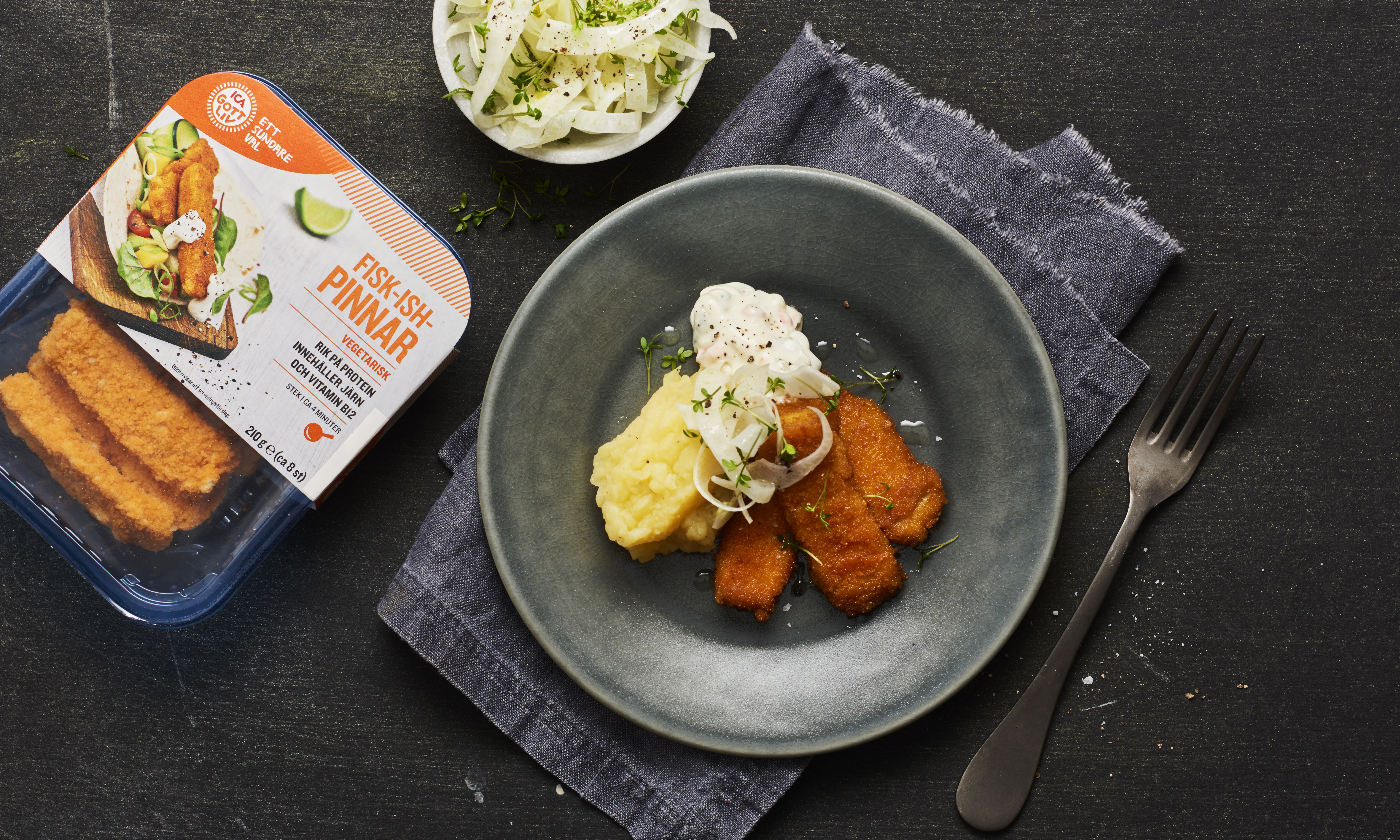 Svart tallrik med veganska fiskpinnar, potatismos och sås bredvid en förpackning veganska fiskpinnar.
