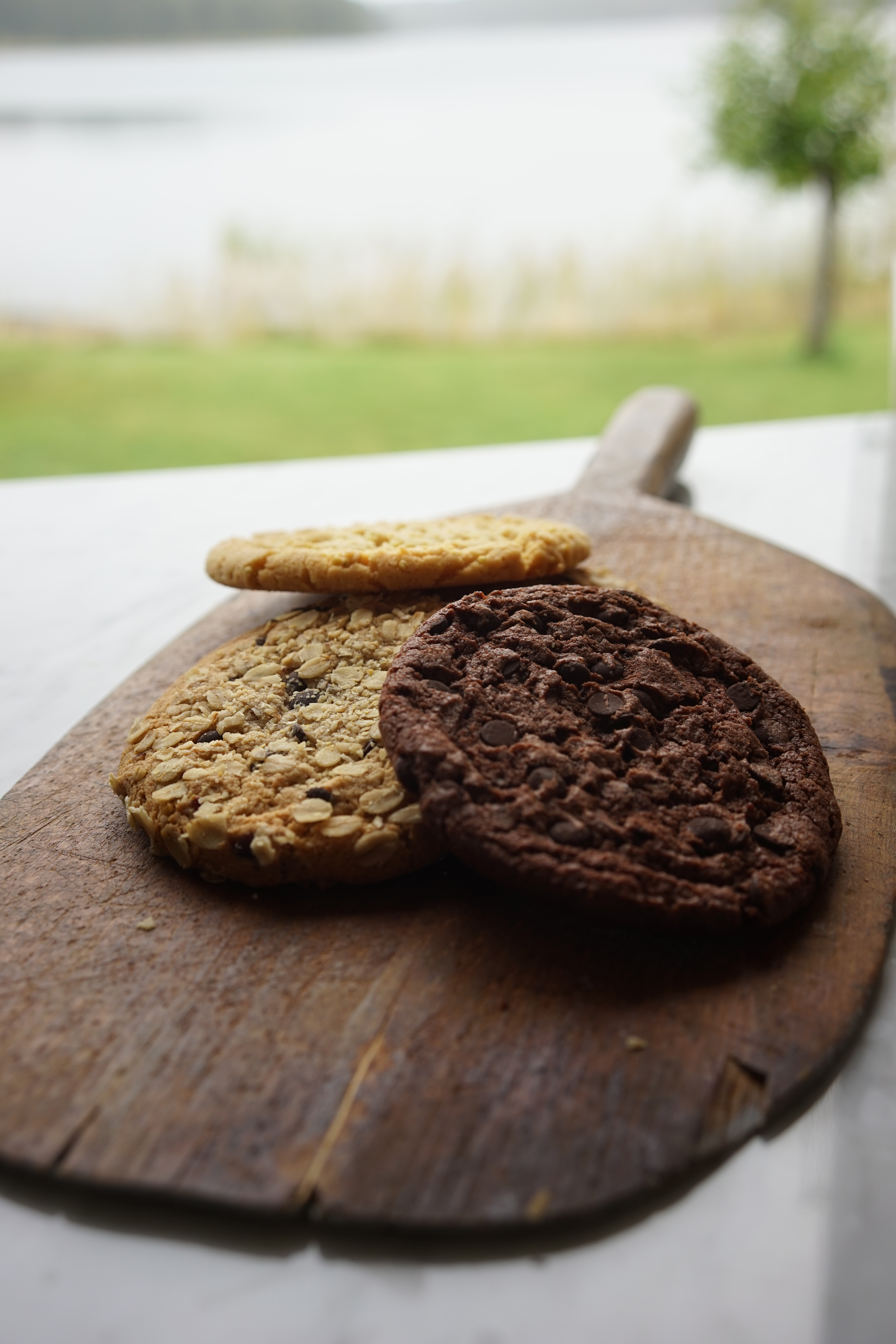 Tre cookies ligger på en skärbräda i trä på ett vitt bord. Två ljusbruna och en mörkbrun.