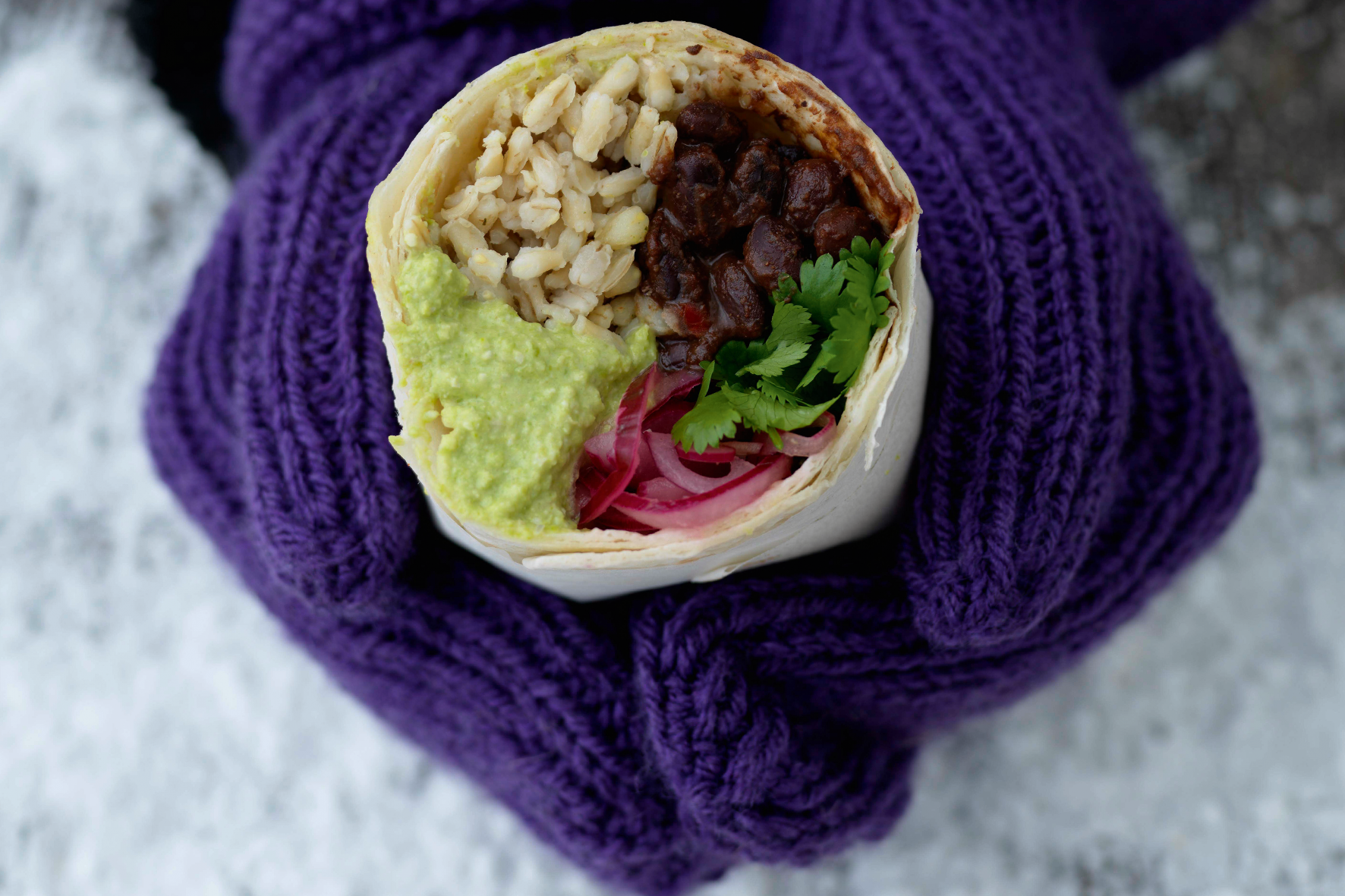 händer med lila vantar håller i en burrrito med krämiga svarta bönor, guacamole och ris