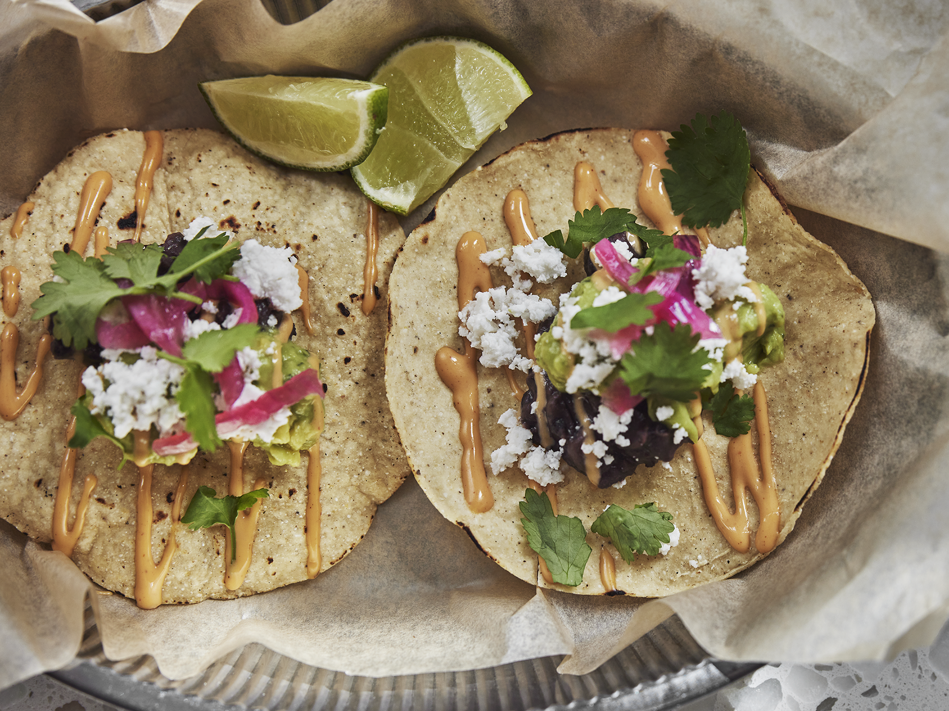 Två, små tacobröd fyllda med bl.a. rödlök, avocado, koriander, vegansk ost och sås, liggandes på bakplåtspapper bredvid limeklyftor.