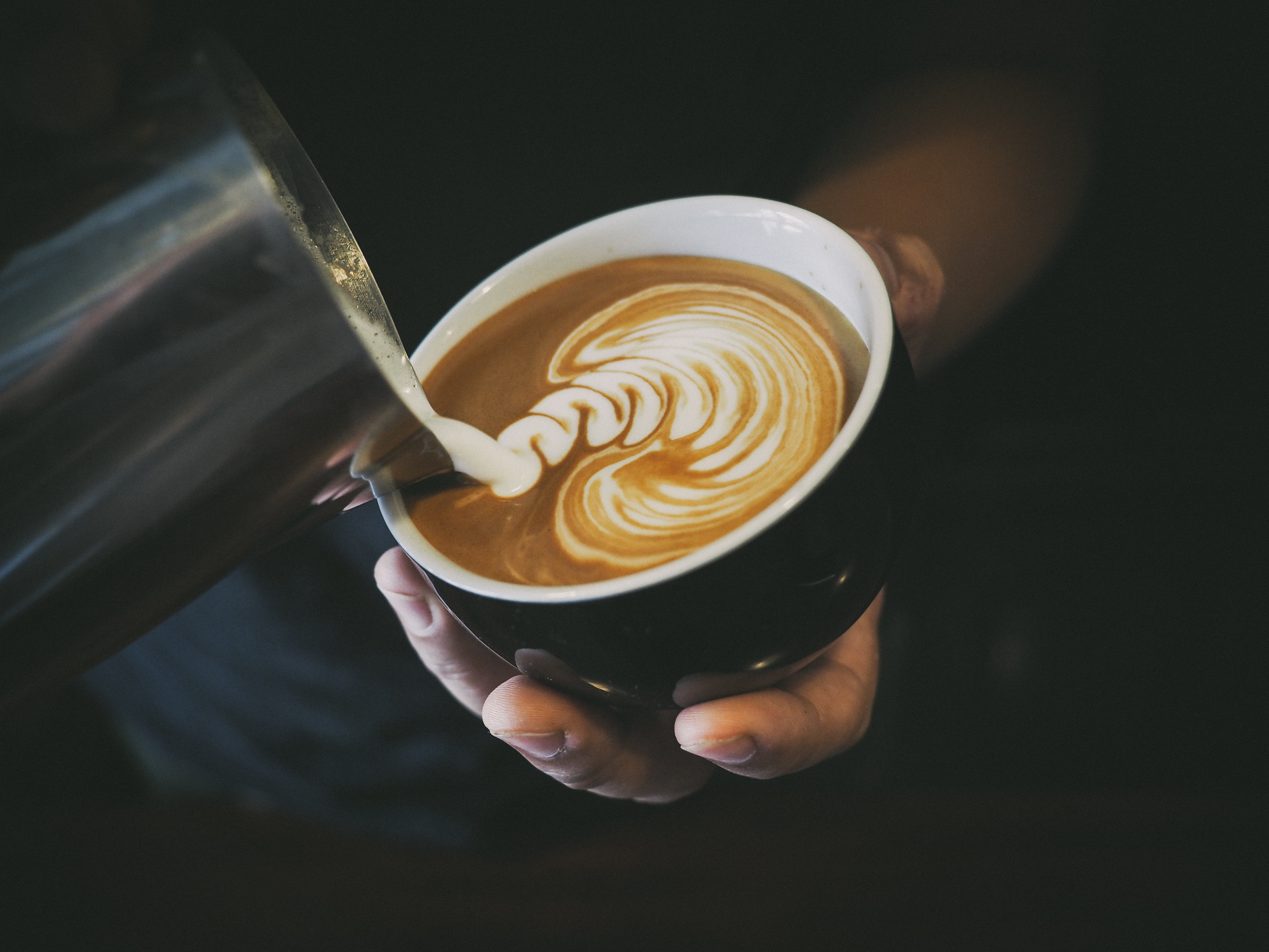 "Mjölk" hälls i kopp med kaffe. Koppen hålls i en hand i dovt belyst miljö.