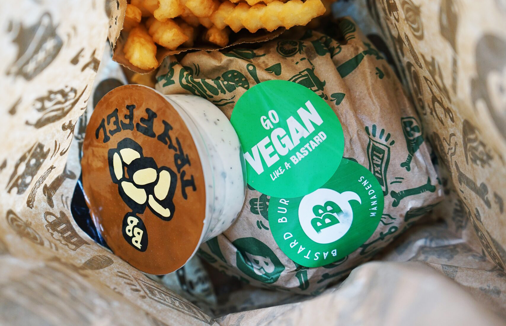 Burgare, pommes och sås från Bastard Burgers i papperspåse. På burgaren finns ett klistermärke med texten "Go vegan like a bastard".