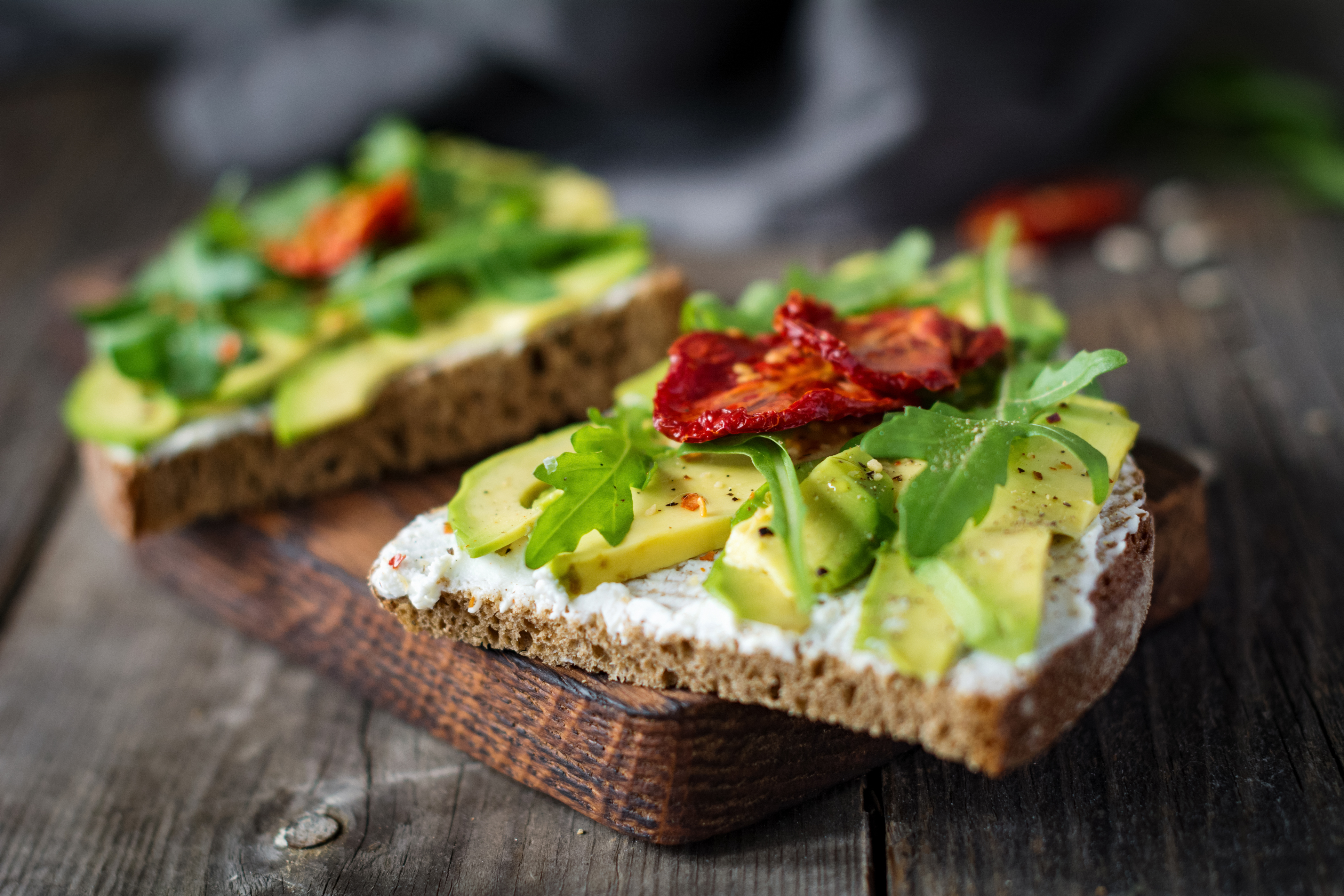 Två rågbrödsmackor med avokado och soltorkade tomater på en skärbräda.