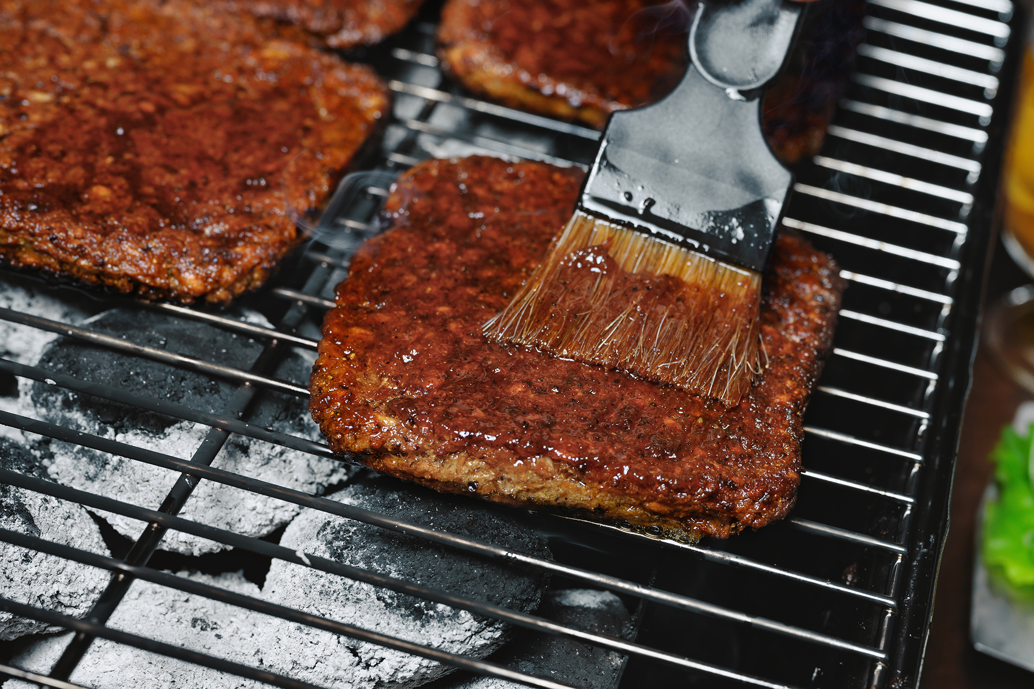 Fyrkantiga veganska burgare på en grill som blir penslad med grillolja.
