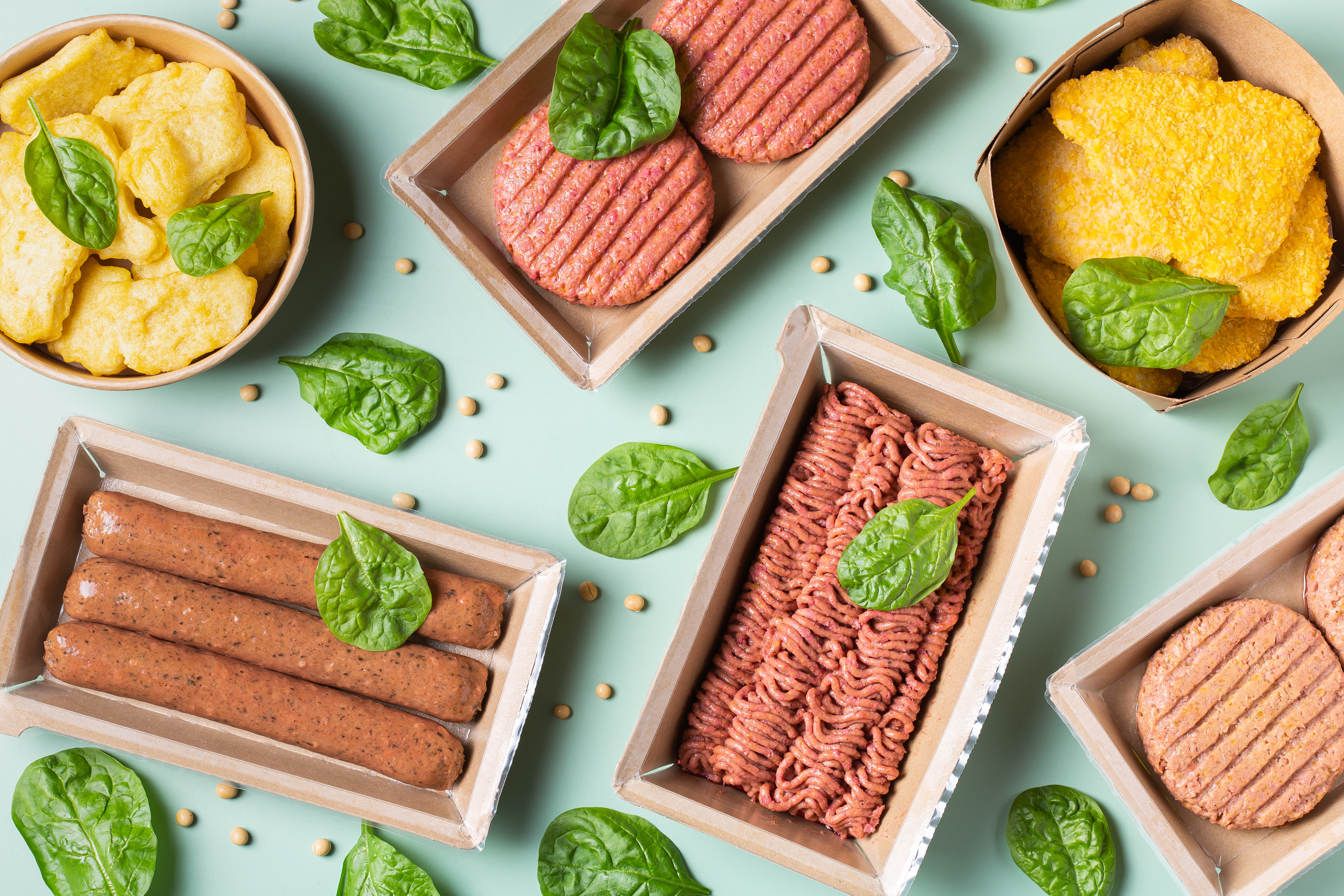 Flera olika växtbaserade köttsubstitut upplagda på ett mintgrönt bord med basilikablad utspridda.
