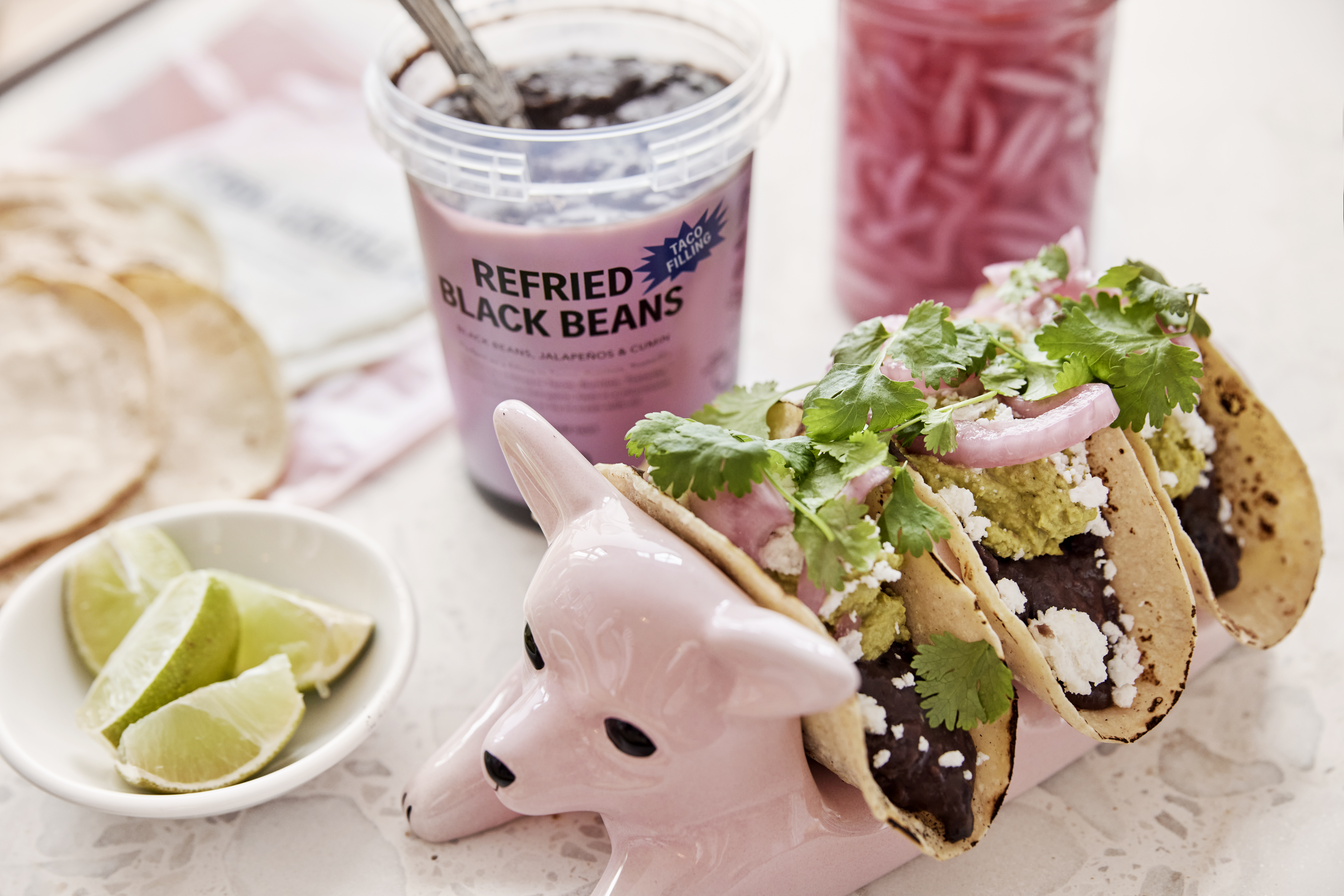 Mjuka fyllda veganska tacos uppställda i en keramikhund med läsk, lime och picklad rödlök på sidan om.