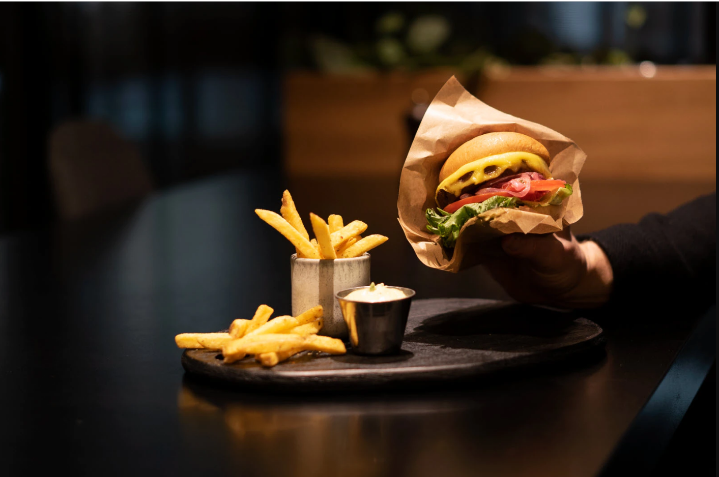 en hand som håller i en vegoburgare med pommes och dip på ett bord i en mörk restaurangmiljö