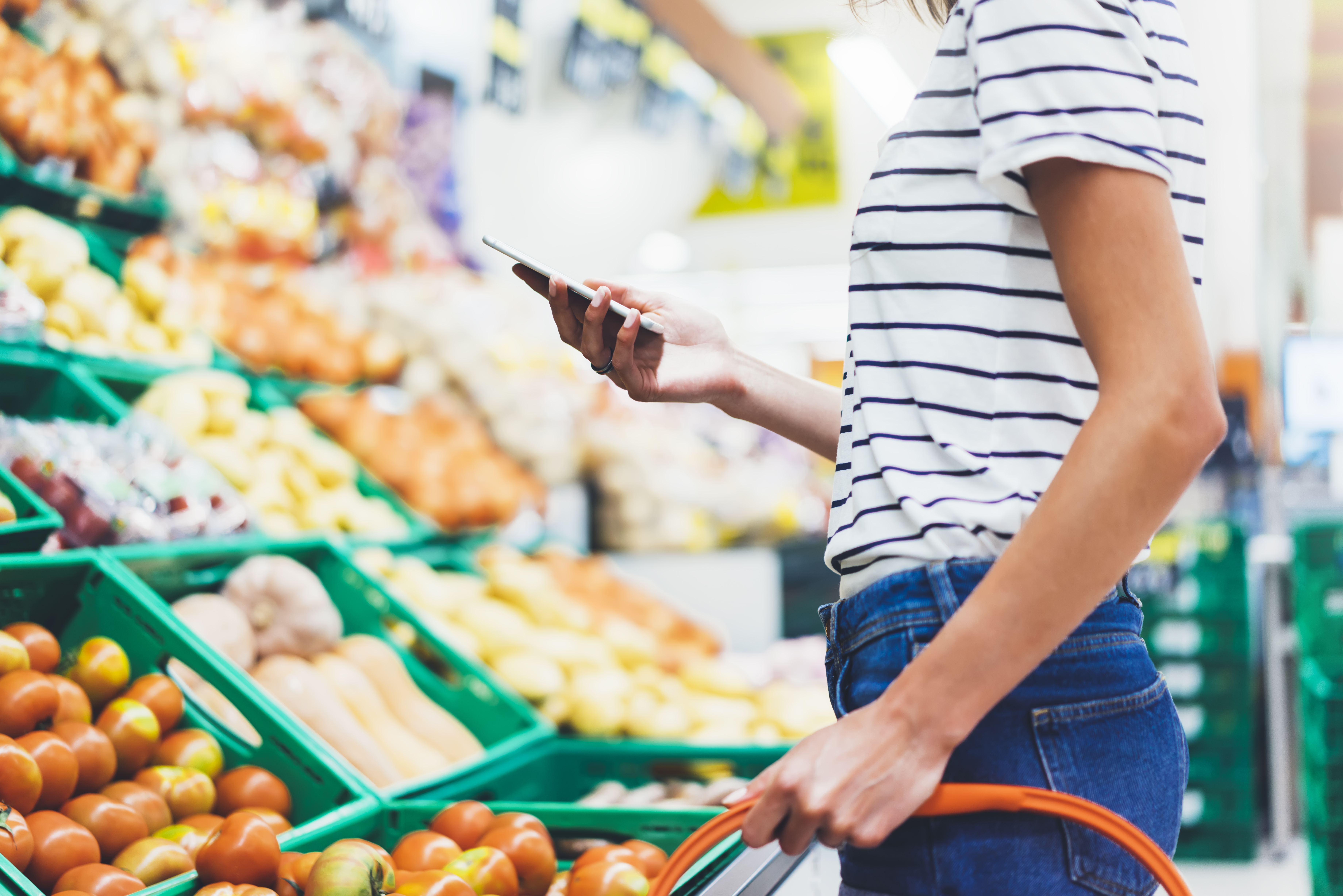 Ung kvinna handlar hälsosam mat i stormarknad suddig bakgrund. Kvinnliga händer köper naturprodukter med smart telefon i butik. Hipster på livsmedelsbutiker med hjälp av mobil. Person som jämför priset på produkter