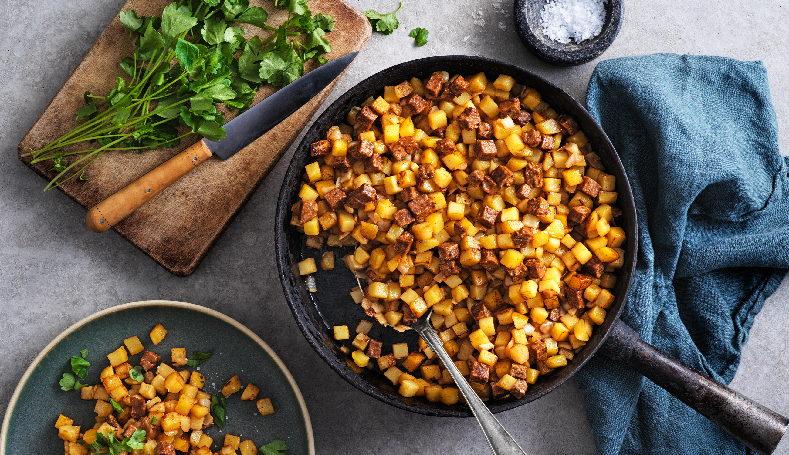 Vegansk pytt i en stekpanna bredvid en tallrik med pytt och en skärbräda med örter