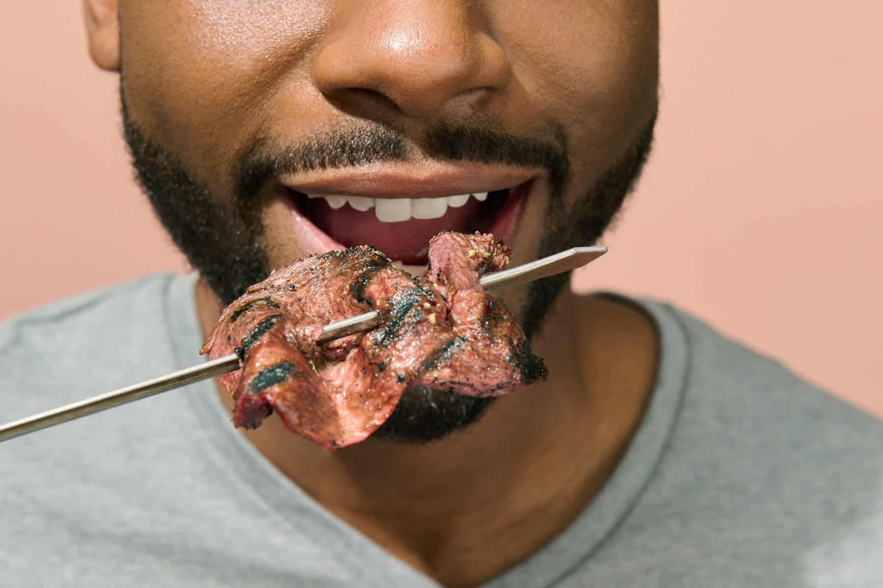 Bild på en mans mun med ett grillspett med kött framför sig