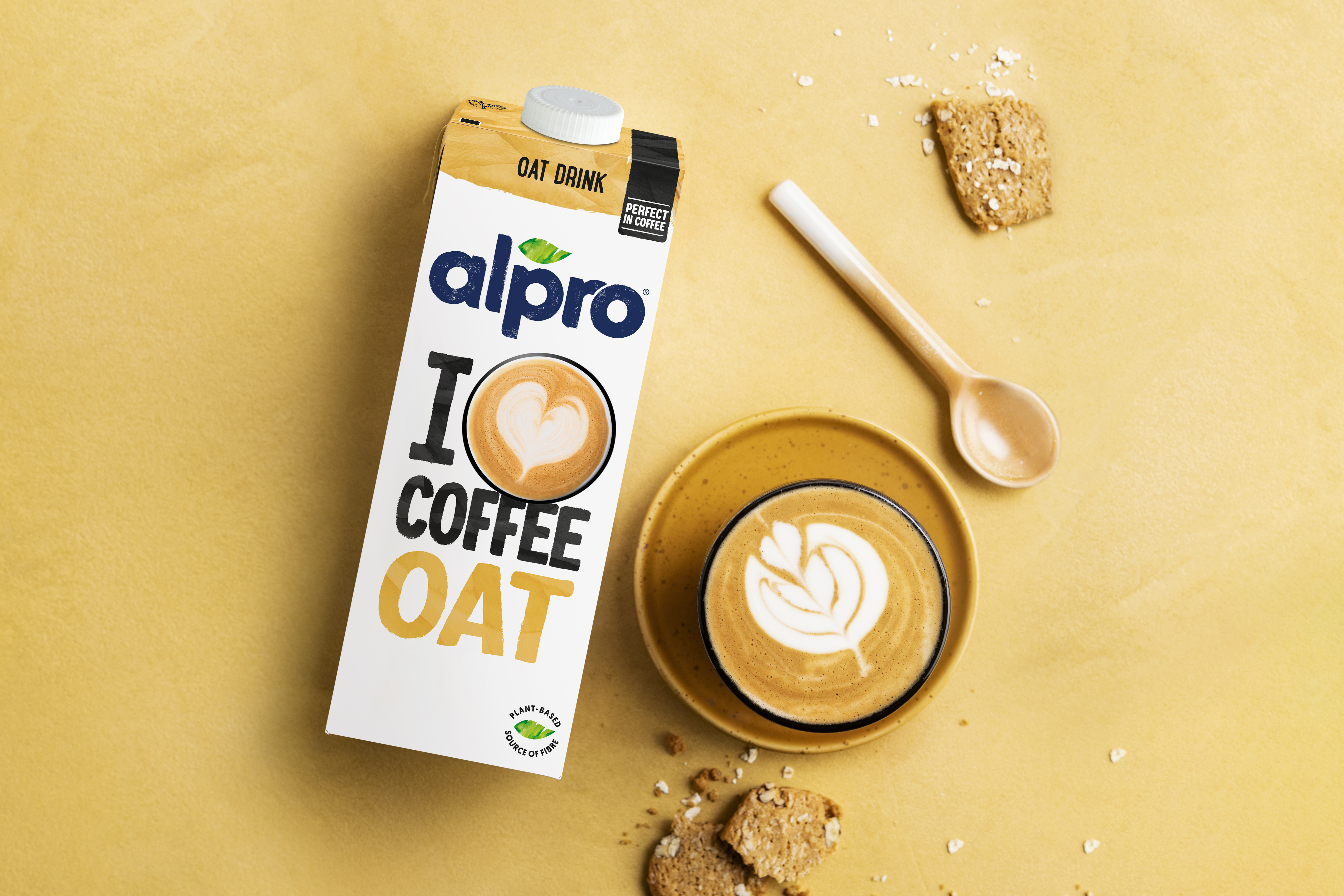 Förpackning med växtbaserad dryck bredvid en kopp latteart på ett gult bord