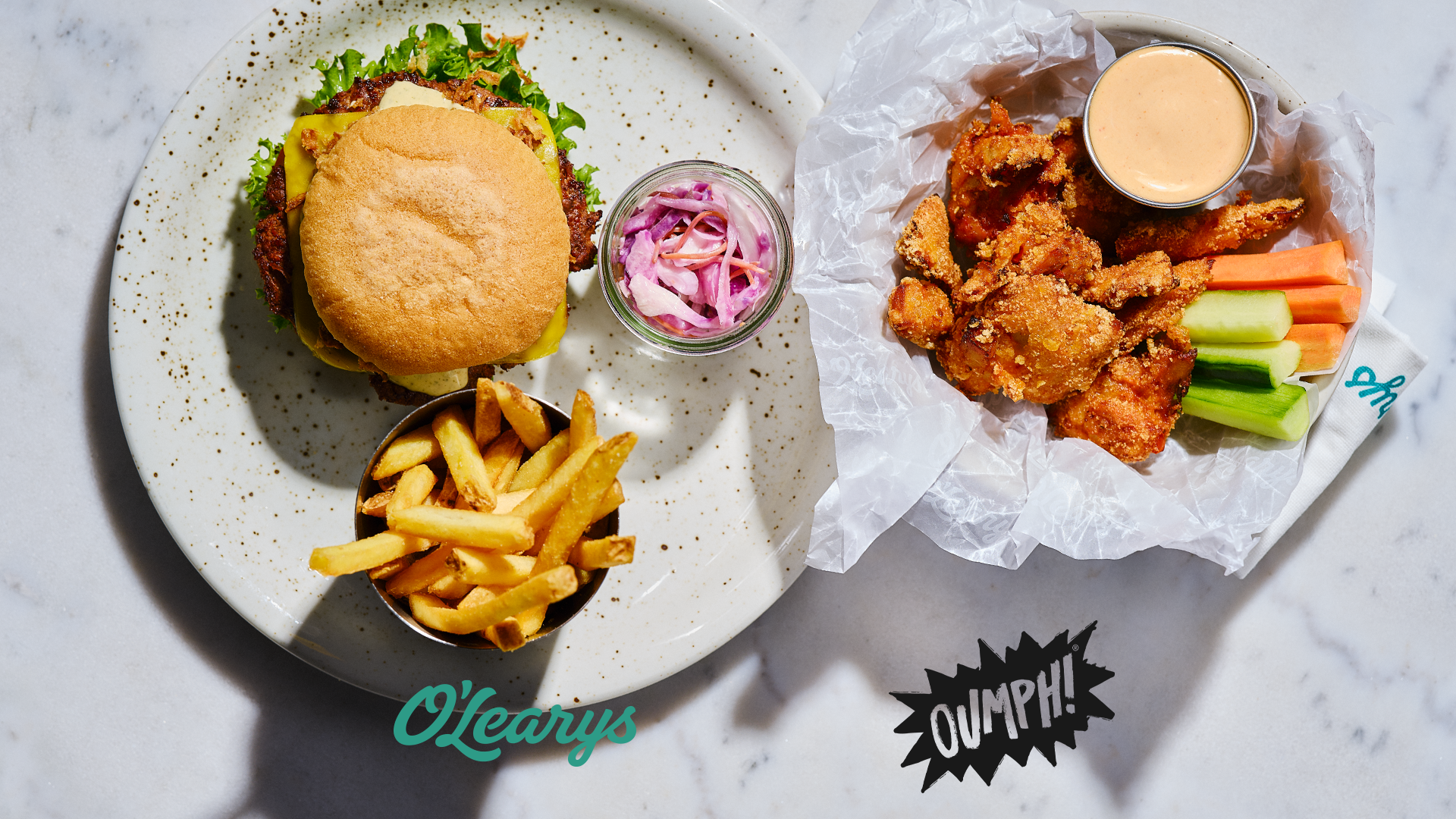 En tallrik med hamburgare, pommes och coleslaw och en med veganska chicken wings, grönsaker och dip. Bild tagen uppifrån.