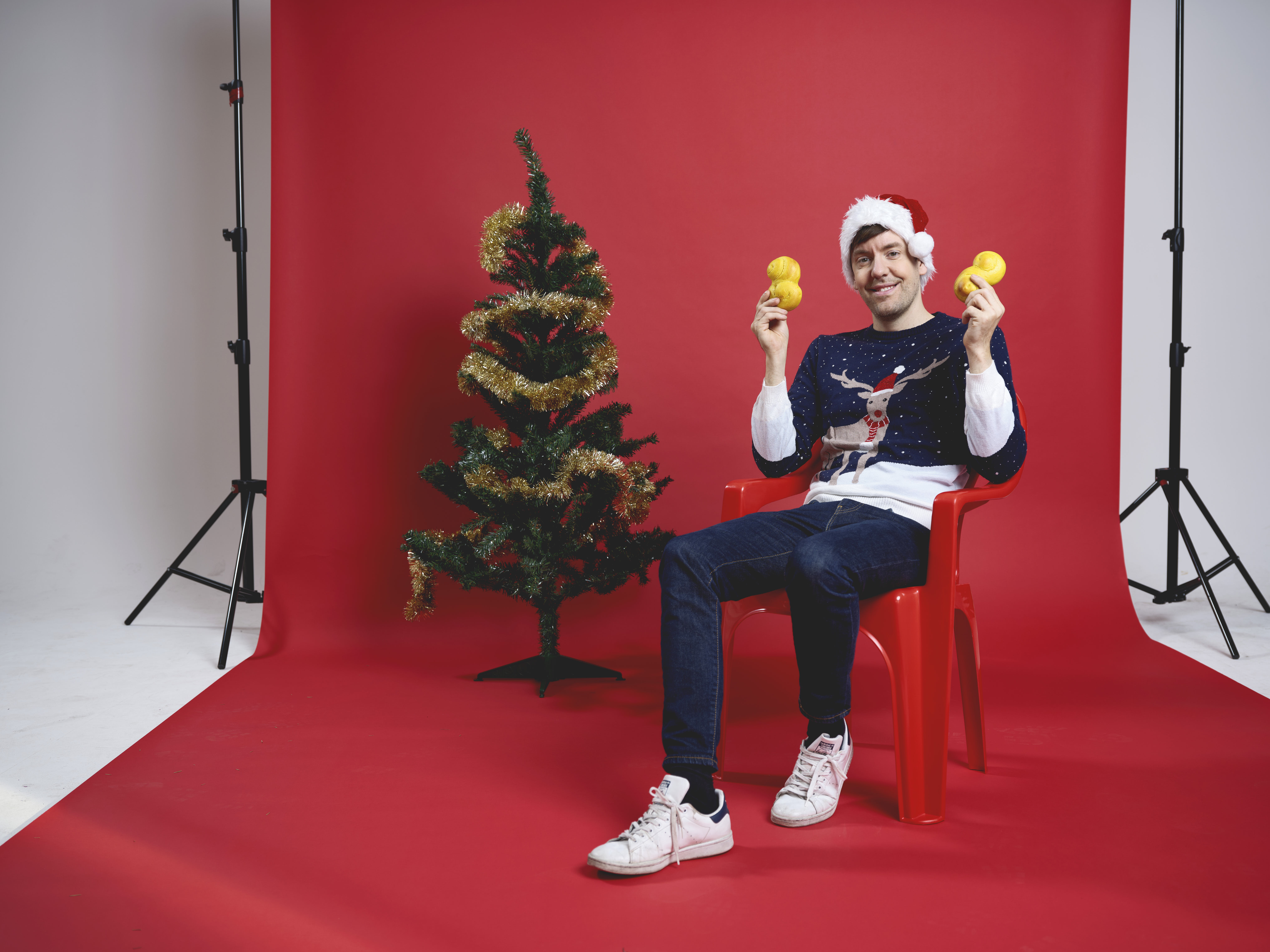 En man som sitter på en stol framför en röd fotovägg. Han håller upp lussebullar, har en jultröja på sig och sitter bredvid en julgran.