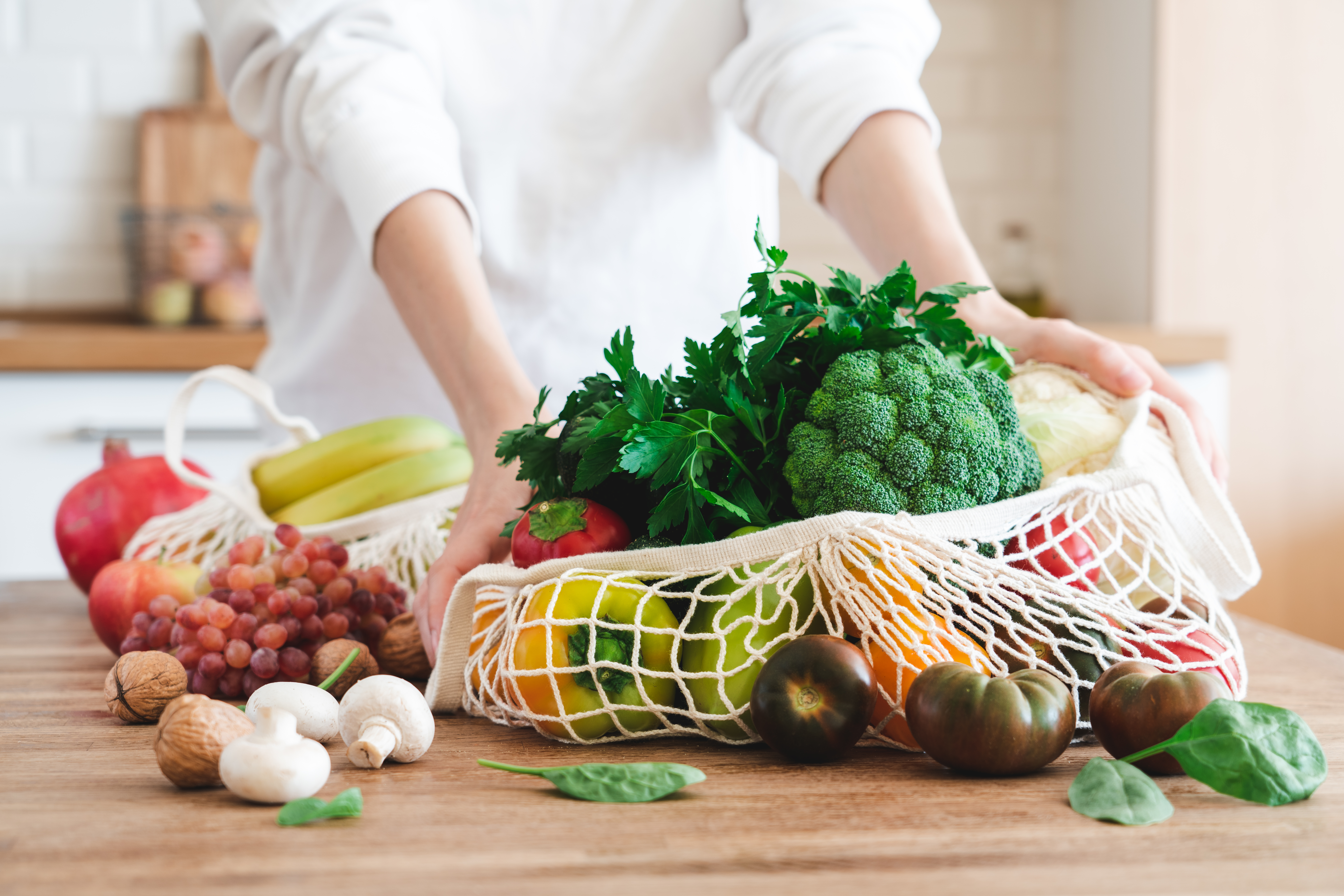 Fruits and vegetables in cotton reusable net mesh bag on wooden table in modern bright kitchen. Organic vegan food set. Sustainable lifestyle concept