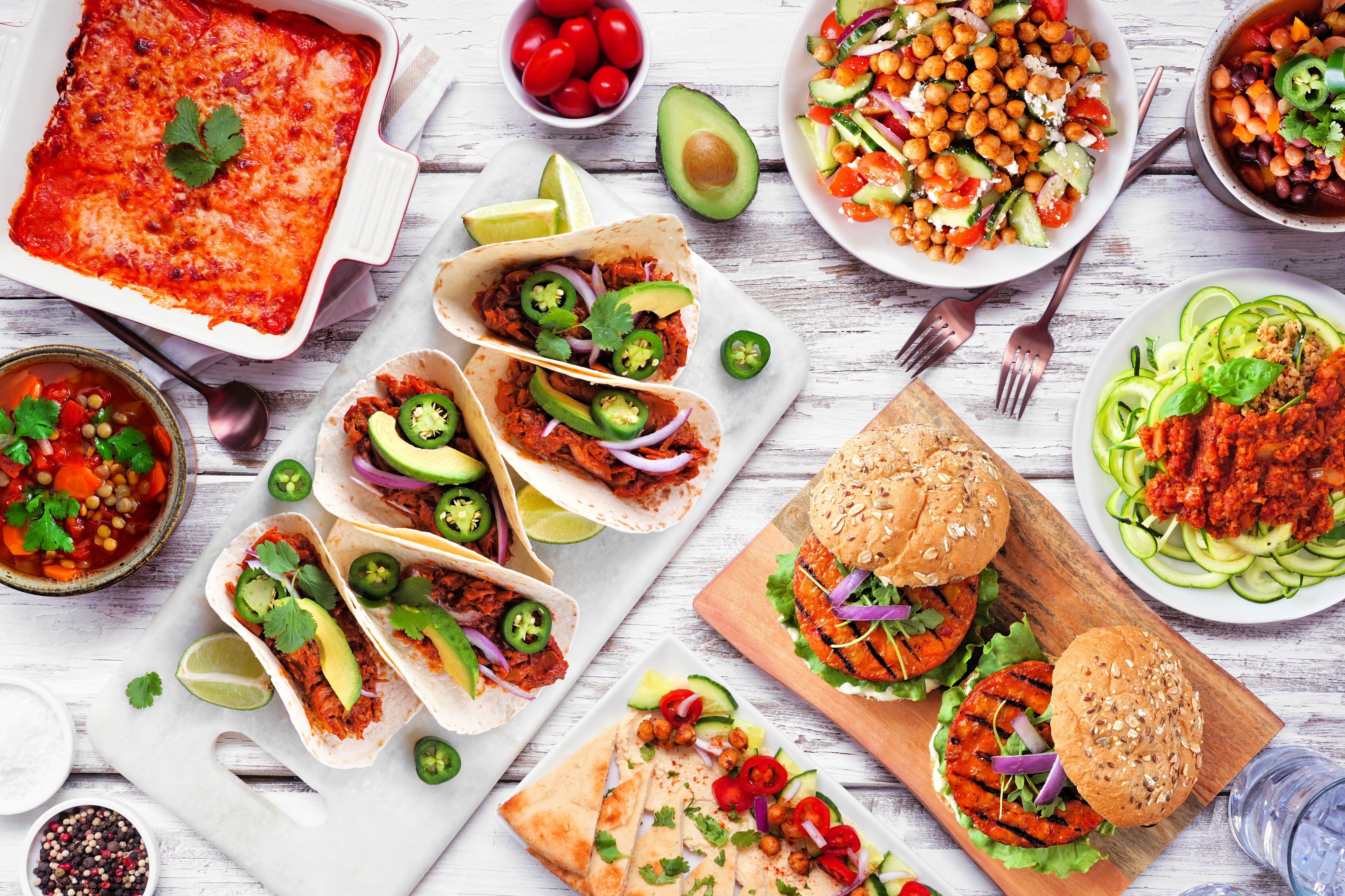 Healthy plant based meal table scene. Above view on a white wood background. Jackfruit tacos, zucchini lasagna, walnut bolognese zoodles, chickpea burgers, hummus, soups, salad.