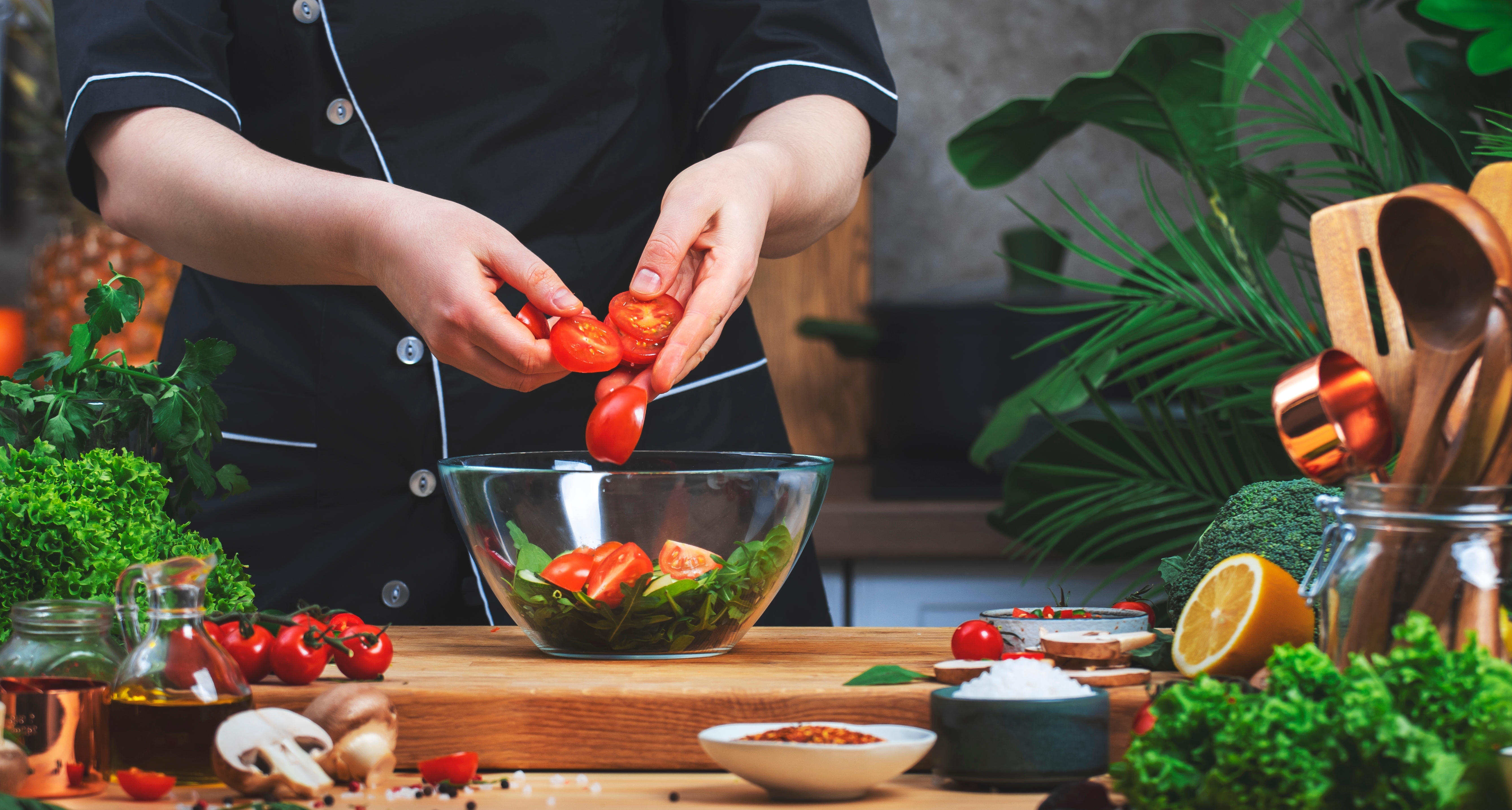 Matlagning i köket. Kocken lägger ​​tomater i skål med ruccola sallad. på träbord med köksredskap, grönsaker, örter och ingredienser. Hälsosamt veganskt ätande, recept, bloggande