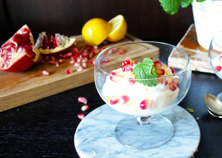 Glasskål med citronmousse och granatäppelkärnor framför en träskiva med granatäpplen och citroner.