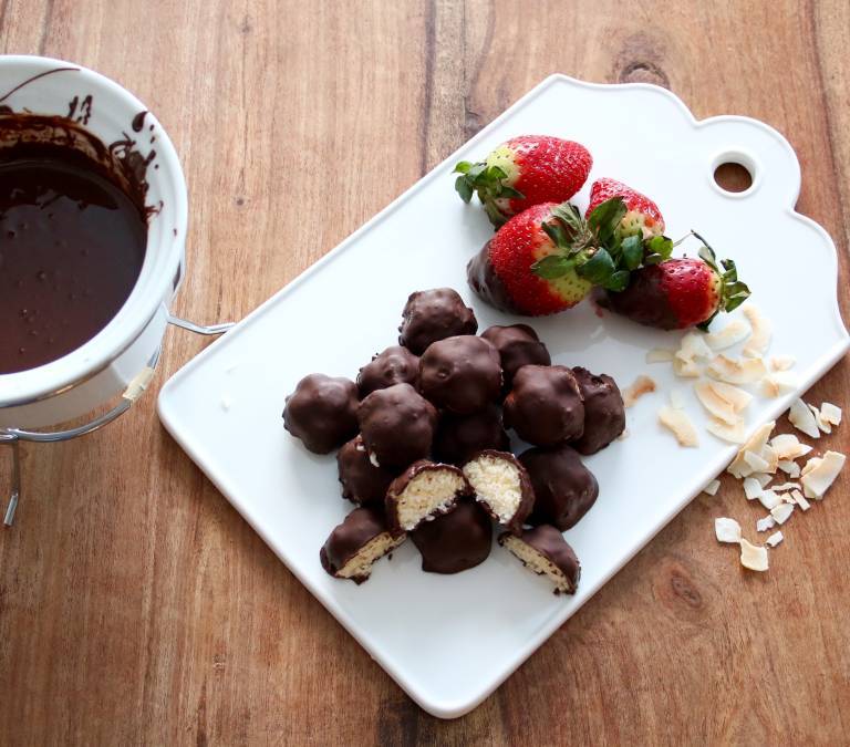 Chokladbitar med kokosfyllning på en vit skärbräda, bredvid ligger några jordgubbar och kokosbitar