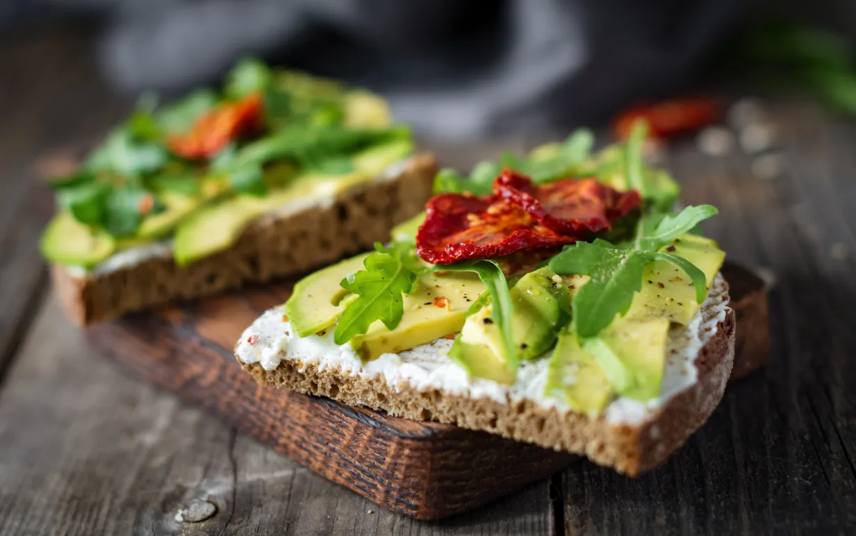 Två rågbrödsmackor med avokado och soltorkade tomater på en skärbräda.