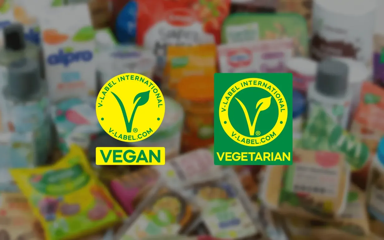 Två uppdaterade V-label loggor mot en suddig bakgrund av livsmedelsprodukter