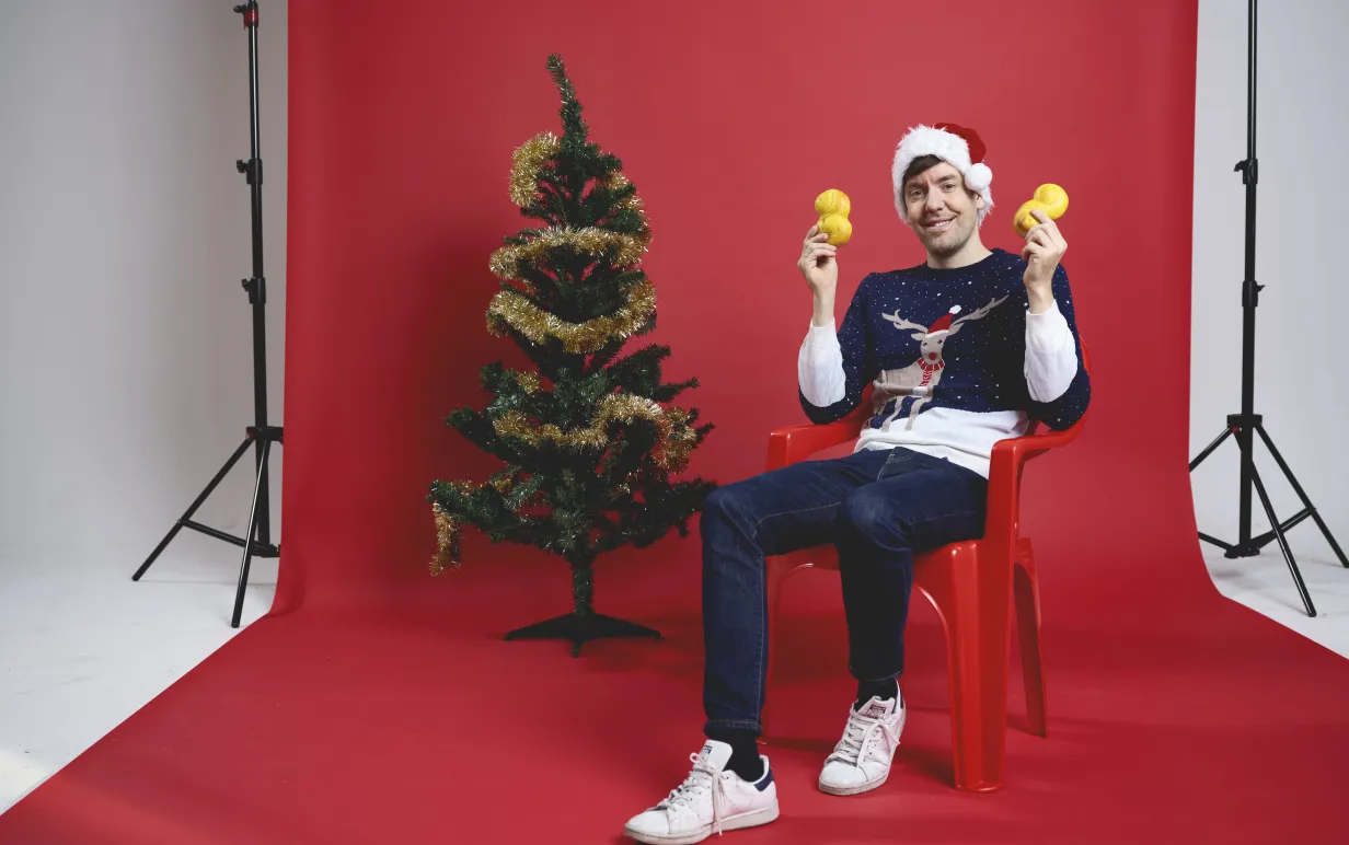 En man som sitter på en stol framför en röd fotovägg. Han håller upp lussebullar, har en jultröja på sig och sitter bredvid en julgran.