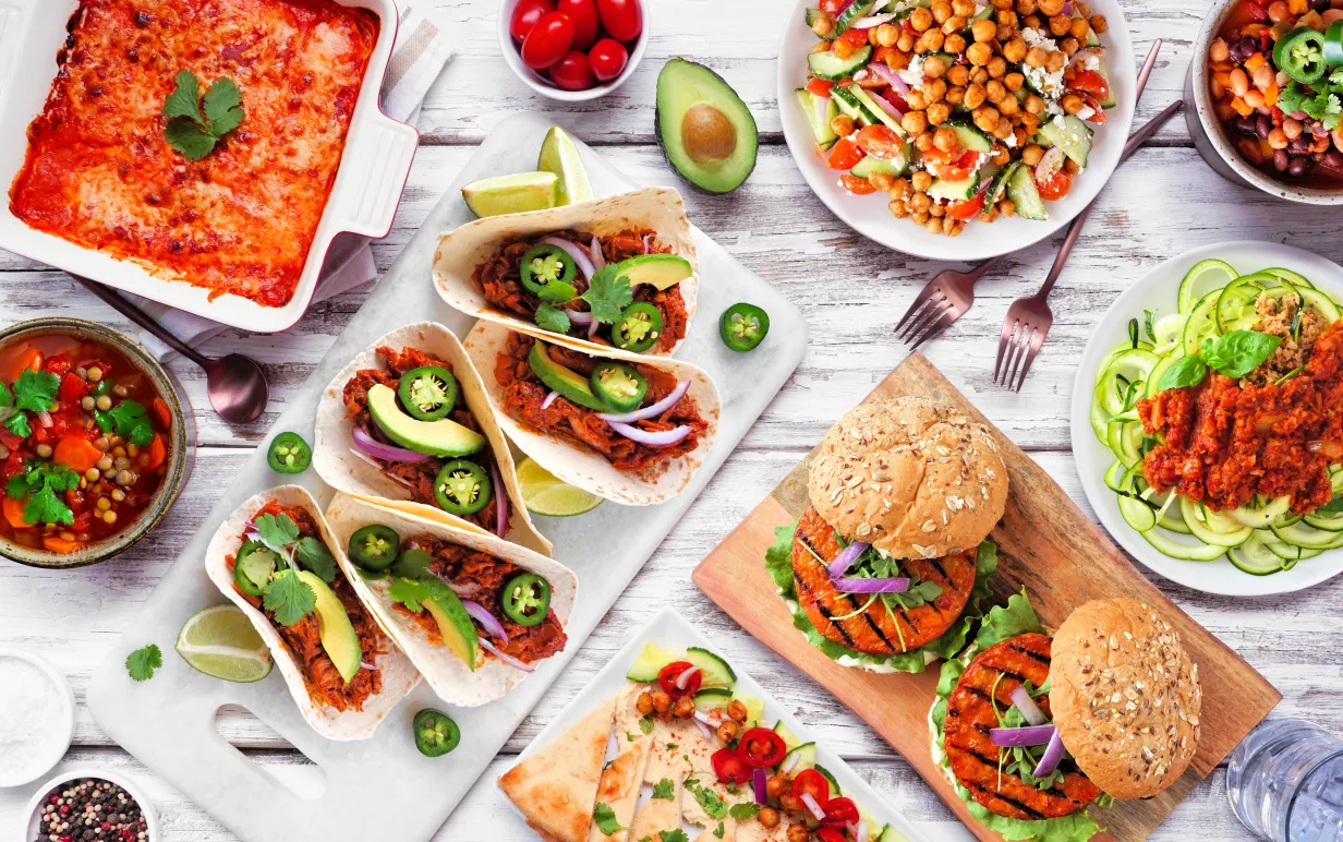 Healthy plant based meal table scene. Above view on a white wood background. Jackfruit tacos, zucchini lasagna, walnut bolognese zoodles, chickpea burgers, hummus, soups, salad.