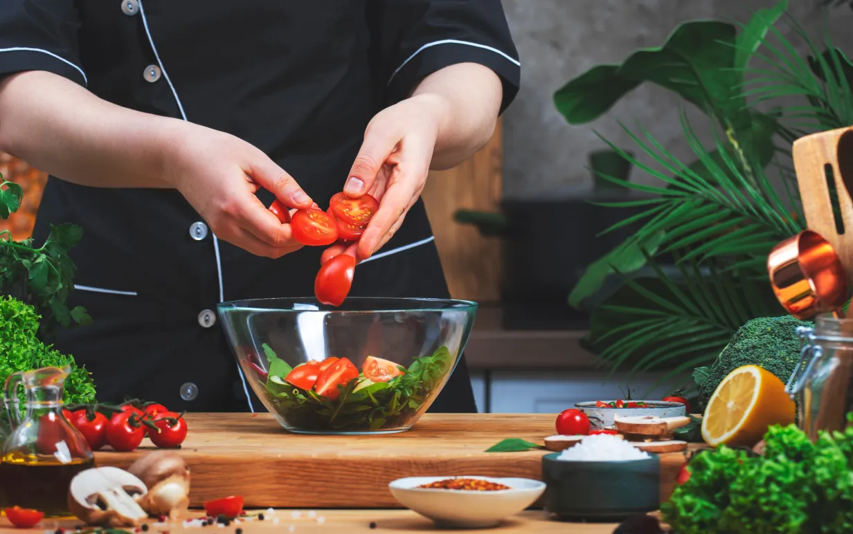 Matlagning i köket. Kocken lägger ​​tomater i skål med ruccola sallad. på träbord med köksredskap, grönsaker, örter och ingredienser. Hälsosamt veganskt ätande, recept, bloggande