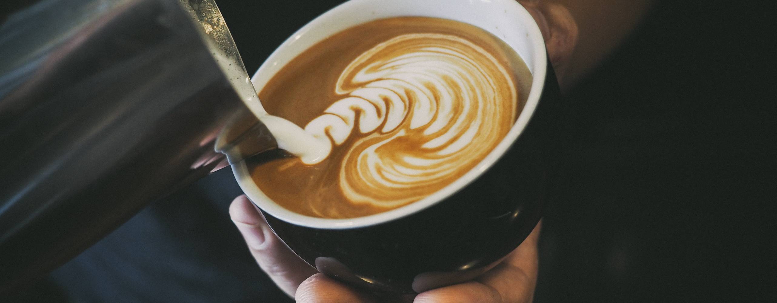 "Mjölk" hälls i kopp med kaffe. Koppen hålls i en hand i dovt belyst miljö.