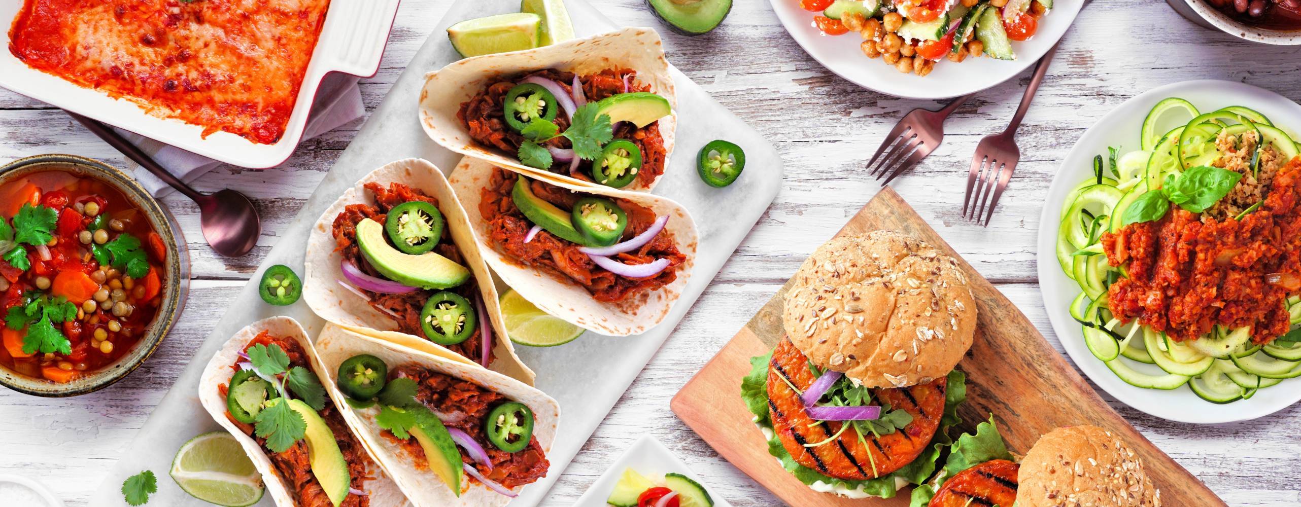Healthy plant based meal table scene. Above view on a white wood background. Jackfruit tacos, zucchini lasagna, walnut bolognese zoodles, chickpea burgers, hummus, soups, salad.